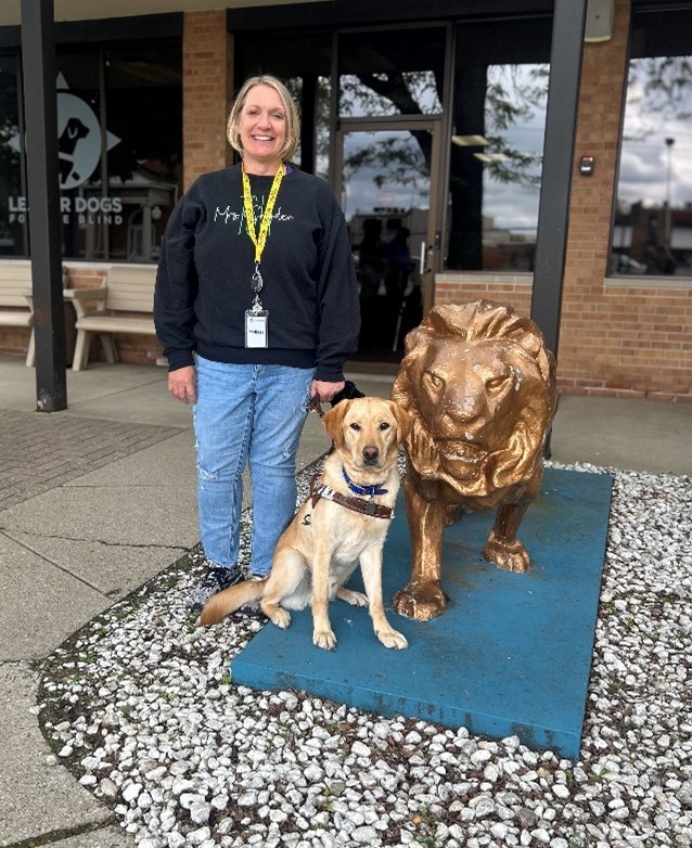 Melissa poses outside the Leader Dogs facility with Viking, a dog guide in training, and a statue of a lion.